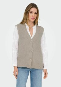 Gilet lavorato a maglia beige con scollo a V e cinque bottoni chiari, indossato sopra una camicia bianca con motivi sottili. Abbinato a jeans azzurri chiari.