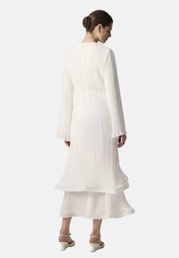 Robe midi blanche plissée avec manches longues, taille cintrée et jupe superposée. Tissu texturé, lien à la taille naturelle, silhouette fluide.