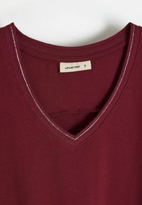 Bordowy T-shirt z dekoltem w serek wykonany z miękkiego materiału, z kontrastującym srebrnym szwem na linii dekoltu oraz wewnętrzną metką wskazującą rozmiar 'S'.