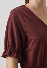 Βουργουνδί φόρεμα με v-λαιμόκοψη και κοντά φουσκωτά μανίκια. Μαλακό, υφασμάτινο ύφασμα με λεπτομέρειες φραμπαλά στις άκρες των μανικιών και με μαζεμένη μέση.