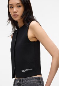 Black sleeveless top with a textured fabric, featuring a round neckline, front buttons, and a white logo "KARL LAGERFELD JEANS" at the hem.