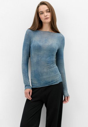 Joven mujer con cabello largo y castaño que lleva una camiseta de manga larga ajustada azul con textura y pantalones negros holgados, de pie contra una pared blanca.