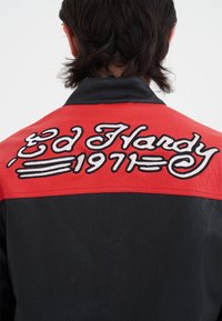 Svart och röd läderjacka med broderad text "Ed Hardy 1971" i vitt på baksidan, med en slät yta och klassisk kragsdesign.