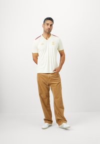 Maglietta a maniche corte crema con dettagli in rosso e oro, caratterizzata da un design a scollo a V e logo ricamato. Abbinata a pantaloni in velluto a coste color sabbia.