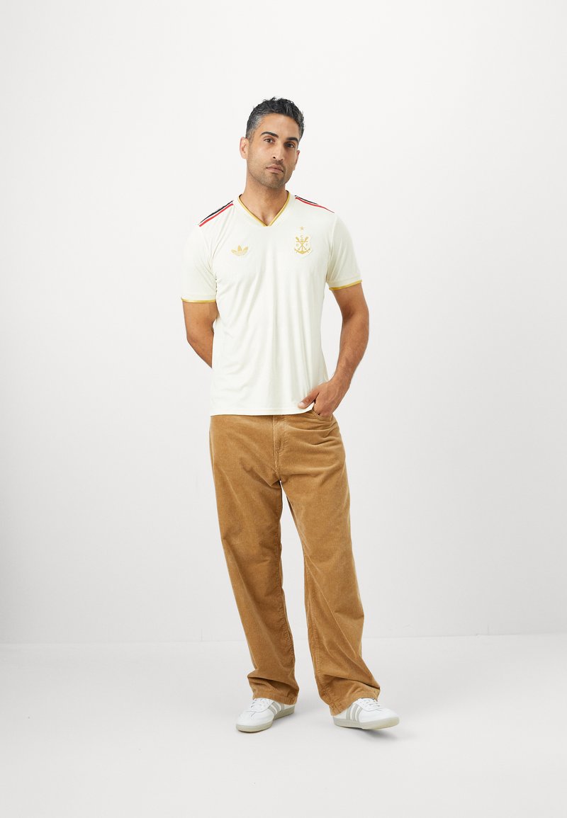 Cremfarbendes Kurzarm-Jersey mit roten und goldenen Akzenten, ausgestattet mit einem V-Ausschnitt und gesticktem Logo. Kombiniert mit beigefarbenen Cordhosen.