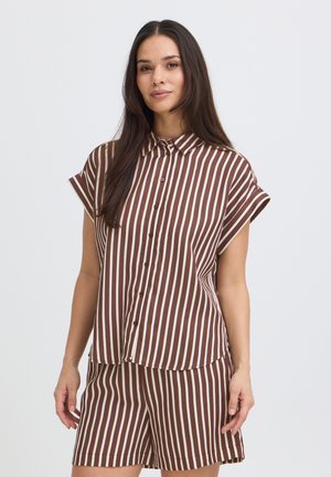 Frau mit langem dunklem Haar, die ein braun-weiß gestreiftes, kurzärmeliges Hemd mit Knöpfen und passende Shorts trägt, steht vor einem einfarbigen Hintergrund.