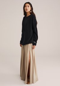 Donna in piedi che indossa un maglione oversized nero, una gonna plissettata beige con uno spacco alto, scarpe marroni e grandi orecchini rotondi su uno sfondo neutro.