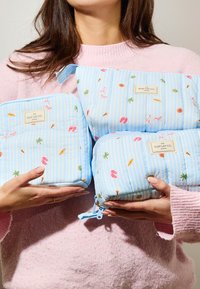 Femme en pull rose tenant trois trousses de voyage bleues décorées de motifs sur le thème de la plage, avec des étiquettes « The Flat Lay Co. London ».