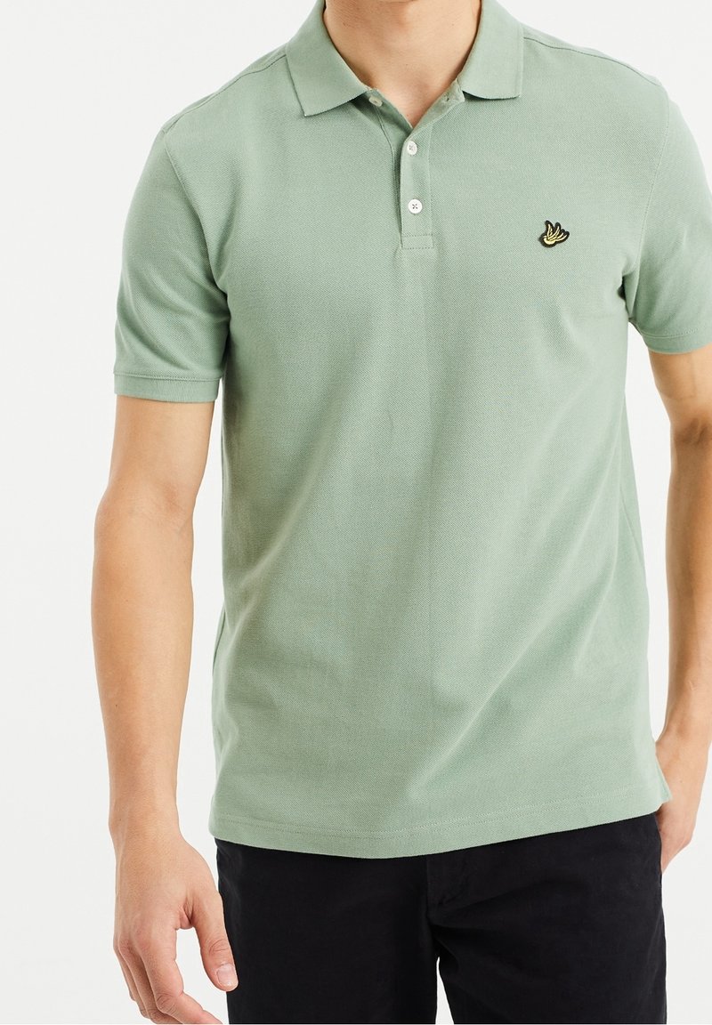Polo en vert clair, en tissu texturé, avec un col à trois boutons et un logo sur la poitrine gauche. Manches courtes.
