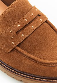 Des mocassins en daim marron présentant une sangle proéminente avec des détails embossés et de petites clous en métal sur le devant. Surface texturée avec une semelle en caoutchouc.