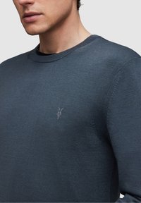 Young man wearing a dark blue knit sweater with a small embroidered animal skull logo on the chest, cropped at the mouth and shoulder.