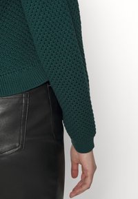 Detailní záběr osoby v texturovaném tmavě zeleném svetru a černých kožených kalhotách, ukazující ruku a paži na bílém pozadí.
