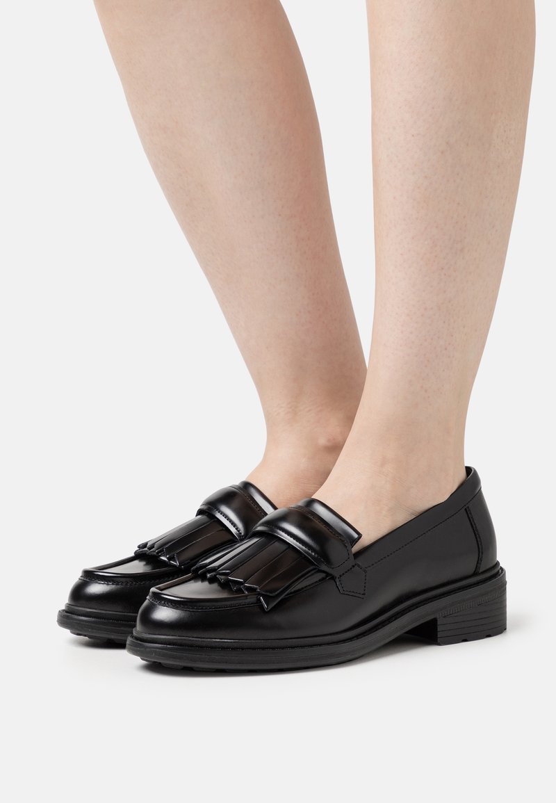 Mocassins en cuir noir avec détail à franges portés pieds nus, vus de profil sur un fond clair uni.
