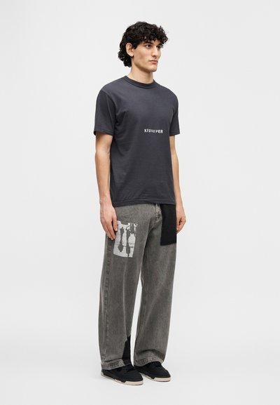 Σκούρο γκρι μπλουζάκι με το "KIDSUPER" τυπωμένο σε λευκό, συνδυασμένο με φαρδιά γκρι τζιν παντελόνια που διαθέτουν αντίθετο  γραφικό κονκάρδα και μαύρες λεπτομέρειες.