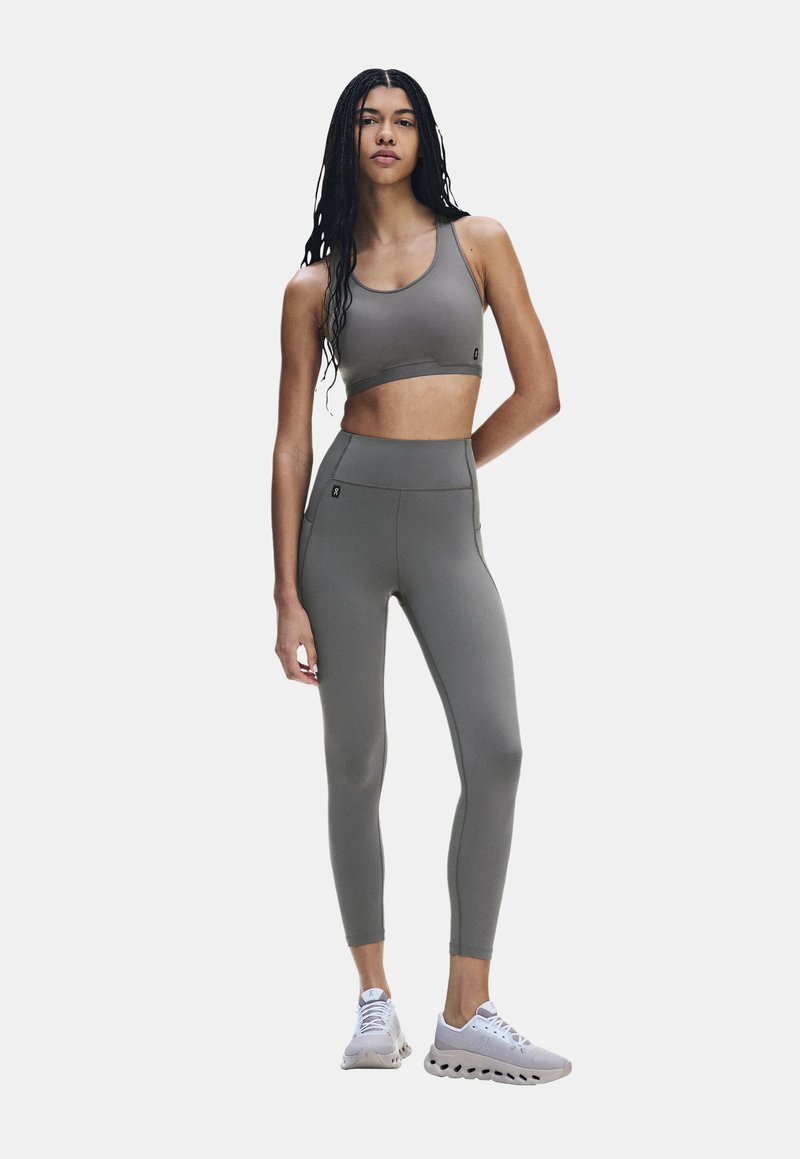 Femme aux longs cheveux tressés portant un soutien-gorge de sport gris, un legging taille haute et des chaussures de sport blanches, debout, une main derrière le dos.