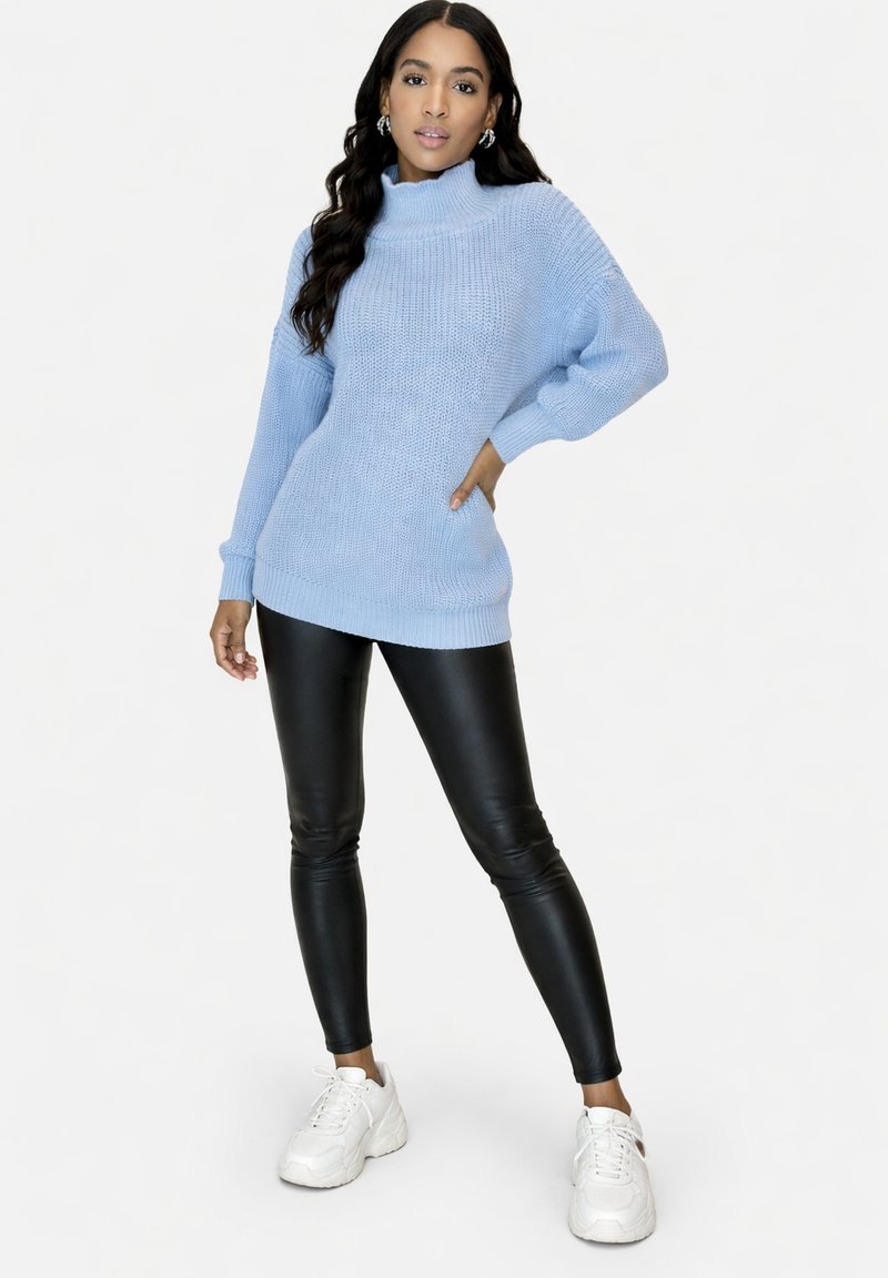 Donna in piedi con la mano sul fianco che indossa un maglione a collo alto azzurro chiaro, leggings in pelle nera e sneakers bianche su sfondo bianco.