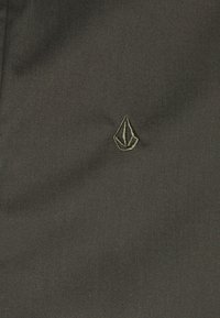 Der schwarze Stoff zeigt ein kleines, goldfarbenes gesticktes Logo in Form eines spitzen Dreiecks. Die Textur ist glatt mit einem leichten Glanz.