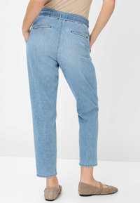 Pantalon en denim bleu clair avec une ceinture extensible, de coupe droite et des poches au dos. Présenté avec des chaussures plates beiges.