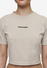 Zkrácená béžová tričko vyrobené z měkké, elastické látky. Obsahuje černě potištěné logo "PEGADOR" na oblasti hrudi. Krátké rukávy, čisté provedení.