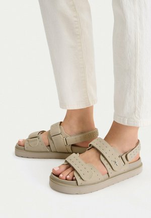 Fødder iført beige flade sandaler med prydede remme og justerbare lukninger, kombineret med off-white bukser mod en ensfarvet baggrund.