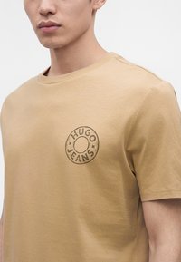 Близък план на бежова тениска с кръгло лого "HUGO JEANS", отпечатано отляво на гърдите, носена от човек с неутрално изражение.