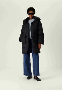 Persona vestida con un abrigo de plumas negro largo, vaqueros de pernera ancha azules, mocasines negros, gafas de sol y un suéter gris claro, de pie sobre un fondo sencillo.