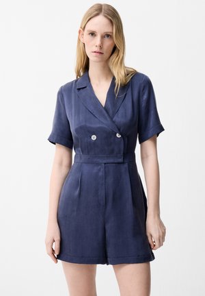 Kvinde med lyst hår iført en mørkeblå kortærmet romper med reverskrave og shorts mod en ensartet lys baggrund.