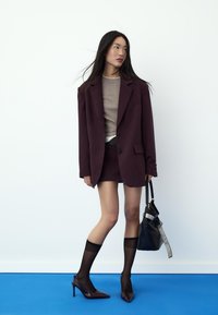 Burgundy tailored blazer over a beige top, paired with a burgundy skirt, black sheer socks, and pointed heels. A black bag with accents completes the look.