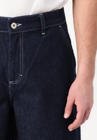 Mørke denimjeans med rette ben, synlige sømmer, en frontlomme og en metalknap på linningen. Hånd i ramme.