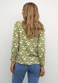 Femme avec de longs cheveux blonds ondulés portant un blouse beige à motifs géométriques verts et un jean en denim bleu, tournée vers l'arrière contre un fond blanc.