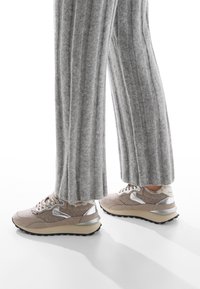 Scarpe da ginnastica beige con una parte superiore testurizzata, dettagli metallici argentati e suola nera robusta, abbinate a pantaloni larghi a coste grigi.