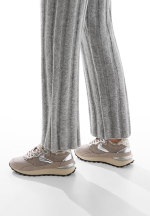 Scarpe da ginnastica beige con una parte superiore testurizzata, dettagli metallici argentati e suola nera robusta, abbinate a pantaloni larghi a coste grigi.