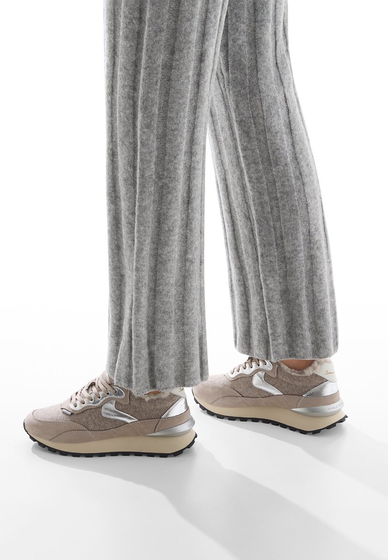 Scarpe da ginnastica beige con una parte superiore testurizzata, dettagli metallici argentati e suola nera robusta, abbinate a pantaloni larghi a coste grigi.