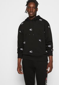 Felpa nera in cotone, con cappuccio regolabile e dettagli logo ripetuti in bianco. Abbinata a pantaloni neri coordinati.