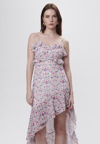 Vestido floral con tirantes finos, cuerpo con volantes y dobladillo asimétrico. Presenta una tela ligera y suave en blanco con patrones en rosa y púrpura.