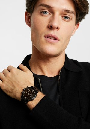 Jeune homme à la peau claire et aux yeux bleus portant une chemise et une veste noires, montrant une montre-bracelet noire à son poignet gauche.