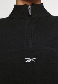 Pull-over noir en polaire avec un col montant et une fermeture éclair à moitié, présentant une surface texturée et un logo blanc sur la poitrine.