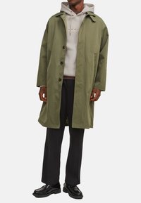 Olivengrüner langer Mantel mit durchgehender Knopfleiste, Reverskragen und großen Seitentaschen, getragen über einem beigen Hoodie und schwarzen Hosen.