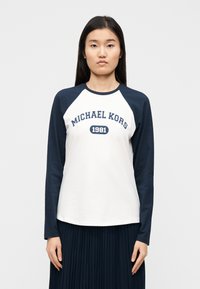 Camisa branca de manga comprida com mangas raglan azul-marinho. Apresenta os dizeres "MICHAEL KORS" e "1981" impressos em azul-marinho na parte da frente. Material macio de algodão.