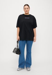 Camiseta negra de gran tamaño con el texto "Day Dreamer", jeans acampanados azules, zapatillas grises y un pequeño bolso negro. Fondo liso.