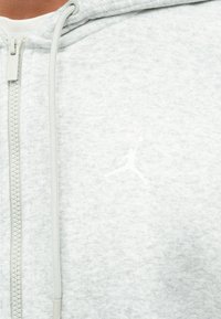 Šviesiai pilkas zip-up džemperis, pagamintas iš tekstūruotos medžiagos, su balta siuvinėta logo ant krūtinės ir atitinkančiomis raišteliais.