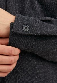 Manica di camicia grigia scura con texture, dotata di un dettaglio con bottone, presenta un piccolo logo e un tessuto morbido e peloso. Inquadratura ravvicinata dell'area del polsino.