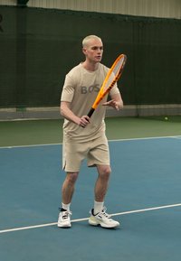Tenista con ropa deportiva beige y el logo "BOSS" se prepara para golpear una pelota. Sostiene una raqueta de tenis naranja en una cancha azul cubierta.
