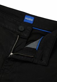 Pantalons noirs avec une étiquette bleue "HUGO", une fermeture éclair à l'avant avec des éléments en métal argenté, et une texture subtile. Poche intérieure en tissu gris.