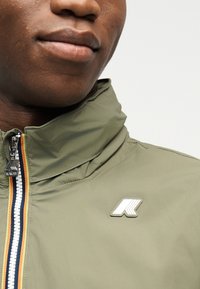 Κοντινό πλάνο ενός ατόμου που φορά μια χακί πράσινη ζακέτα με πολύχρωμο ριγέ φερμουάρ και λευκό λογότυπο στο στήθος.