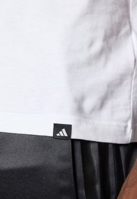Witte katoenen t-shirt met een zwart Adidas-logo label aan de zoom; gecombineerd met donkere, textuurstof broeken met verticale strepen.