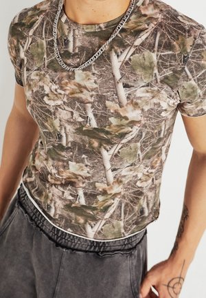 Ein Mann trägt ein enganliegendes T-Shirt mit Camouflage-Muster, eine silberne Halskette, schwarze Hosen mit Kordelzug und hat ein Tattoo am rechten Unterarm.