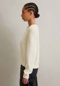 Cremfarbener, gestrickter Pullover mit strukturiertem Finish, der einen Rundhalsausschnitt und gerippte Bündchen aufweist, kombiniert mit schwarzen, hochgeschnittenen Hosen.