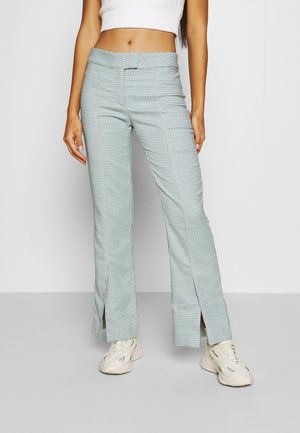 Kvinde iført højttaljede blå og hvide hundetandsmønstrede bukser med frontslids og hvide sneakers, stående mod en ensartet lys baggrund.