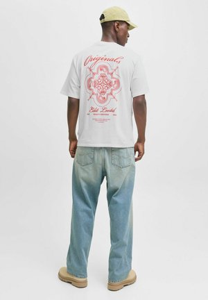 Hvid t-shirt med rød grafisk design på ryggen, der har udsmykkede detaljer og tekst. Løse lyseblå jeans og beige fodtøj fuldender looket.